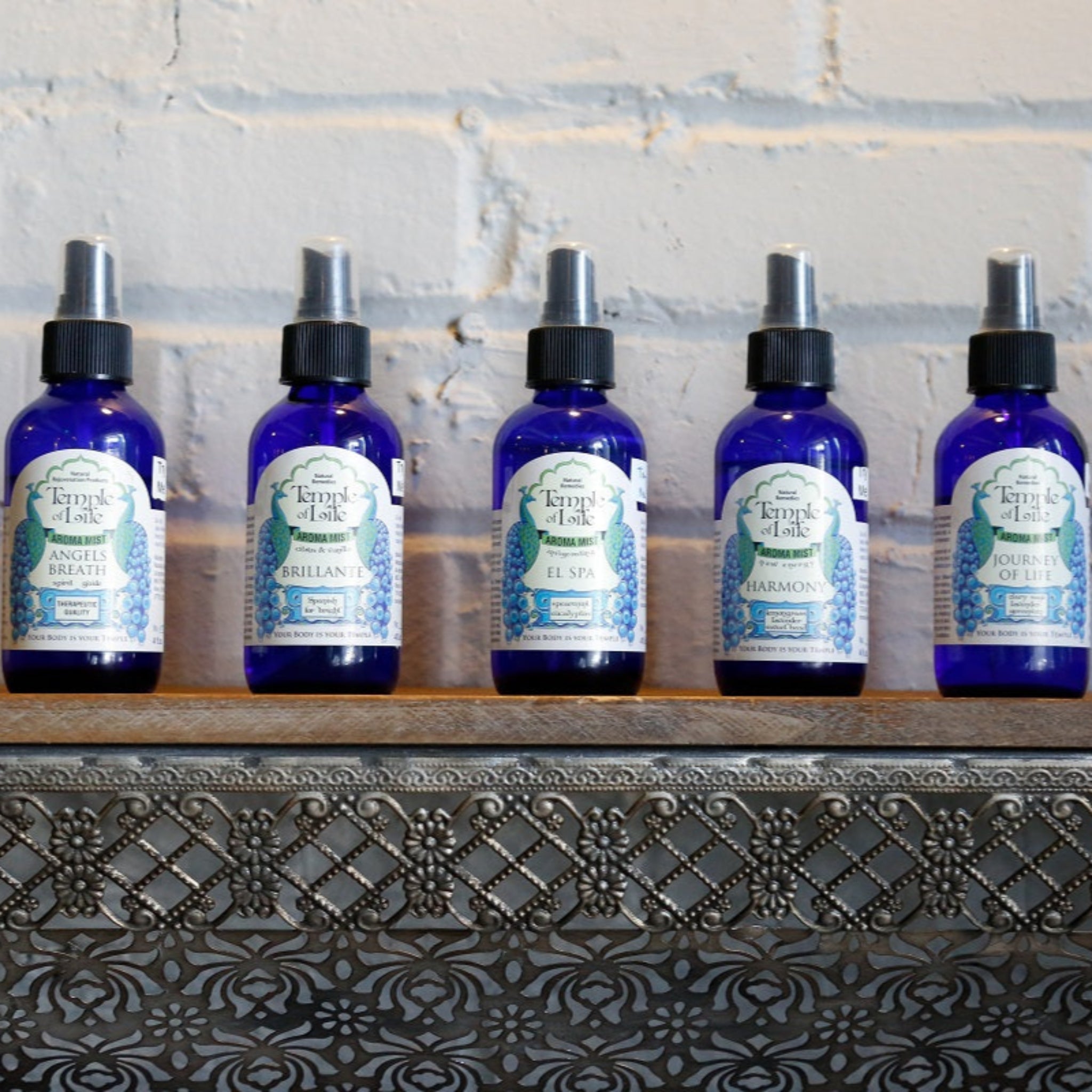 Six blue bottles of natural room sprays with labels on a decorative shelf against a stone wall.